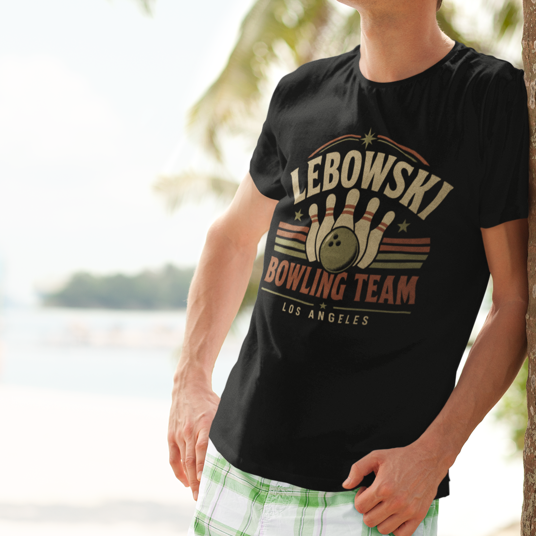 A man wearing a black graphic t-shirt with a retro bowling team design, leaning against a palm tree on a sunny beach.