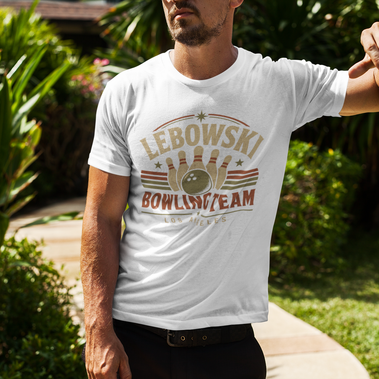 A man wearing a white graphic t-shirt with a 'Lebowski Bowling Team Los Angeles' design, leaning against a palm tree in a sunny, tropical garden setting.