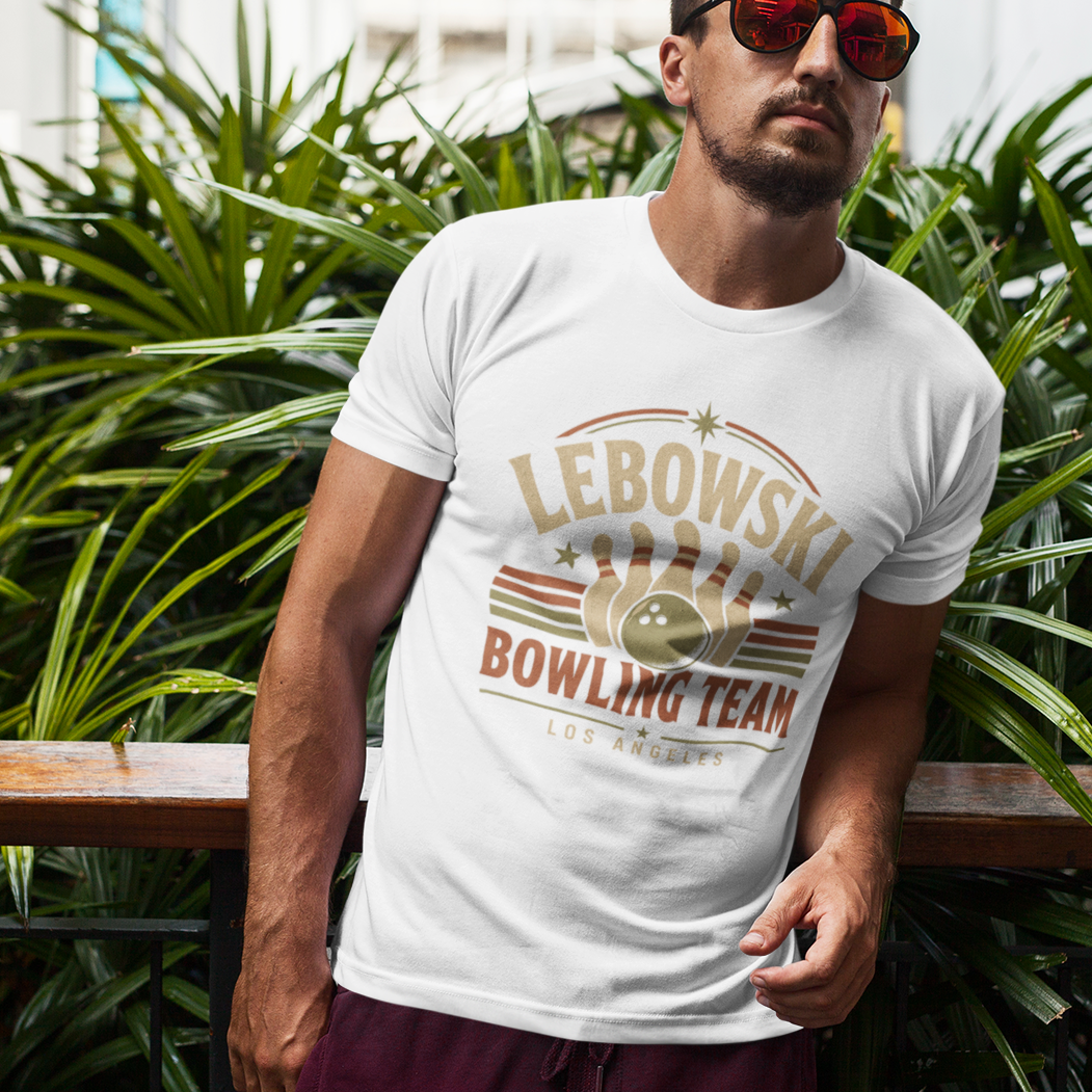 A man wearing a white graphic t-shirt with a 'Lebowski Bowling Team' design, standing outdoors in front of lush green plants.